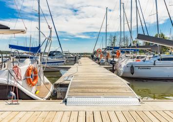 Pomost z odnogami cumowniczymi. Przy y-boomach przycumowane łódki. W tle błękitne niebo z białymi chmurkami.
