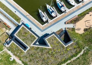 Na pierwszym planie budynek kapitanatu z lotu ptaka. Zarysowują się geometryczne kształty nieregularnej bryły budynku wbudowanego w skarpę i przykrytego trawą. Przy nadbrzeżu przycumowane łodzie żaglowe.