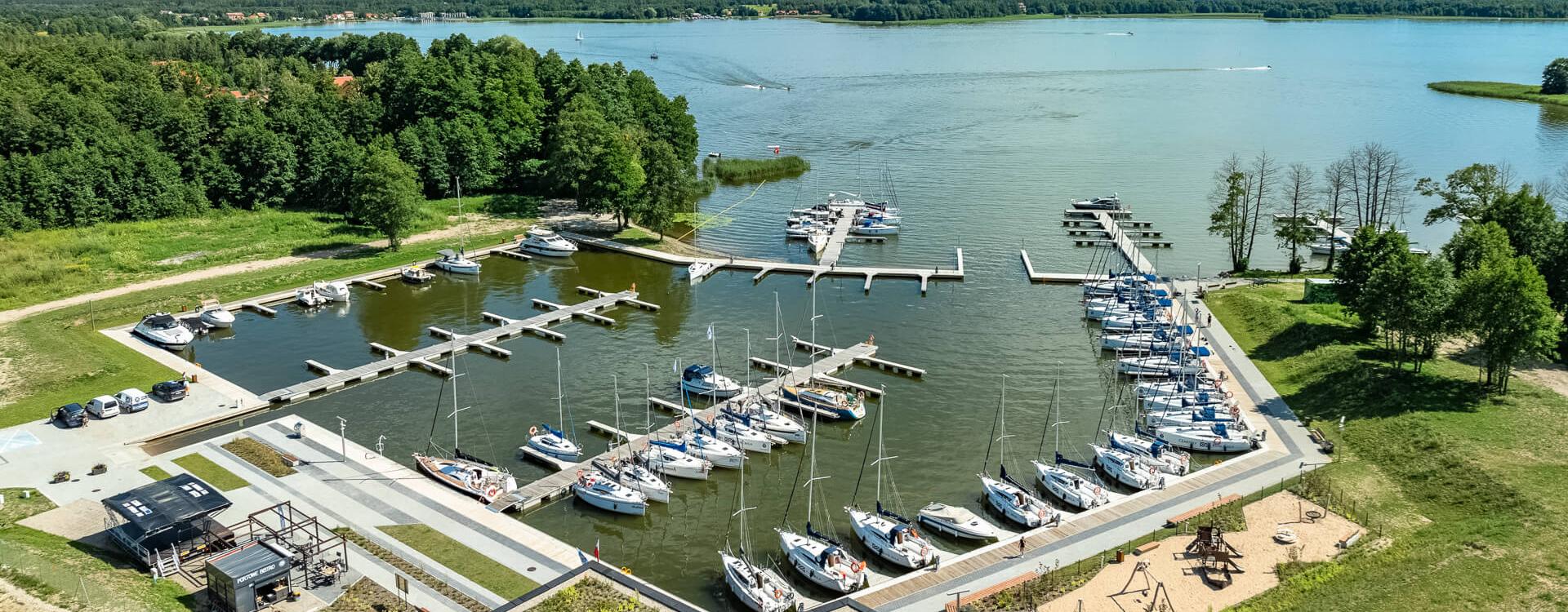 Widok na Przystań Nautica z lotu ptaka. Na pierwszy planie pawilon kapitanatu, budynek wbudowany w skarpę i przykryty trawą. Po prawej stronie keje czarterowe. Na środku basen portowy i wejście do portu. Z lewej u góry widać keje rezydenckie. W tle widać jezioro Święcajty 